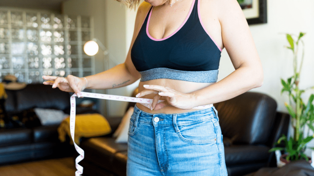 Woman measuring her waist with a tape measure at home while wearing a sports bra and jeans, tracking weight loss progress in a casual living room setting.