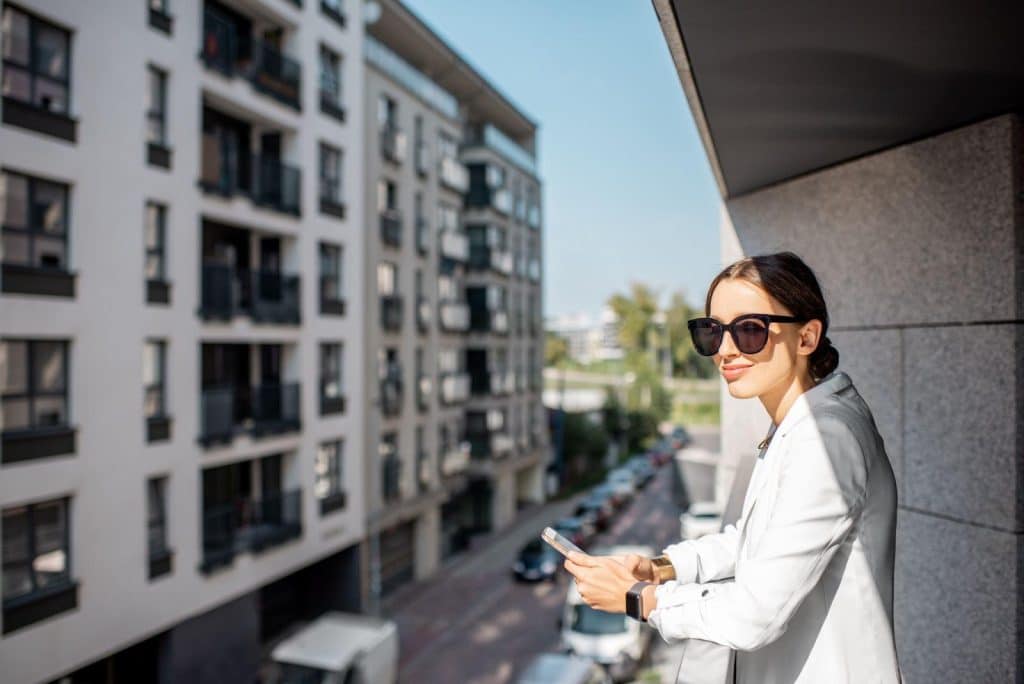 Young professional standing on modern apartment balcony in Dallas, overlooking residential buildings with phone in hand)